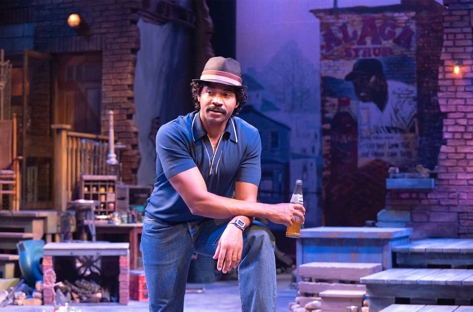 A performer in a brimmed hat leans on one knee while holding a glass bottle. The surrounding brick courtyard and scattered household objects suggest a neighborhood gathering place.