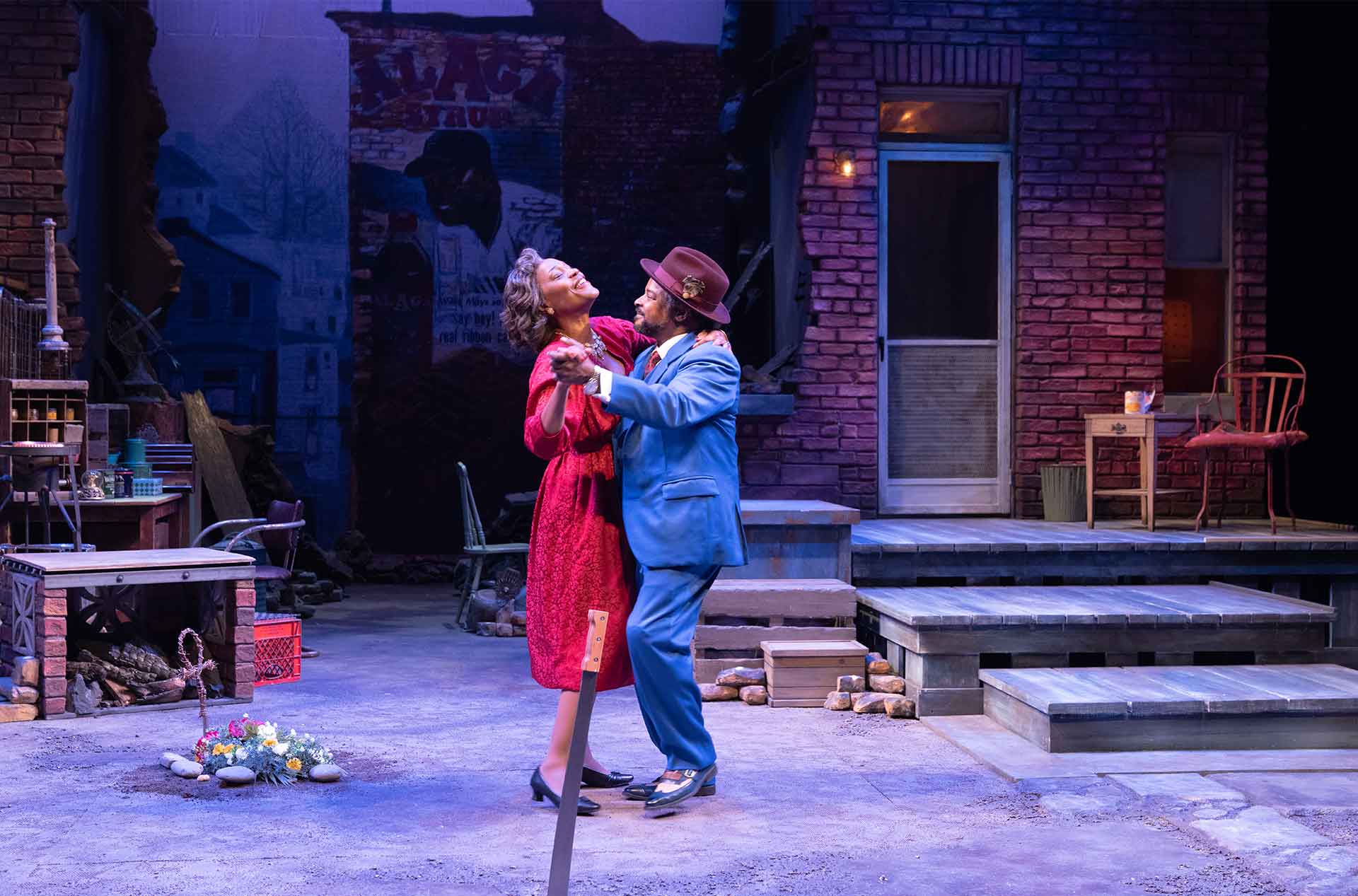 Two performers dance closely in the center of a brick courtyard set. One wears a red dress while the other wears a bright blue suit and brown hat.