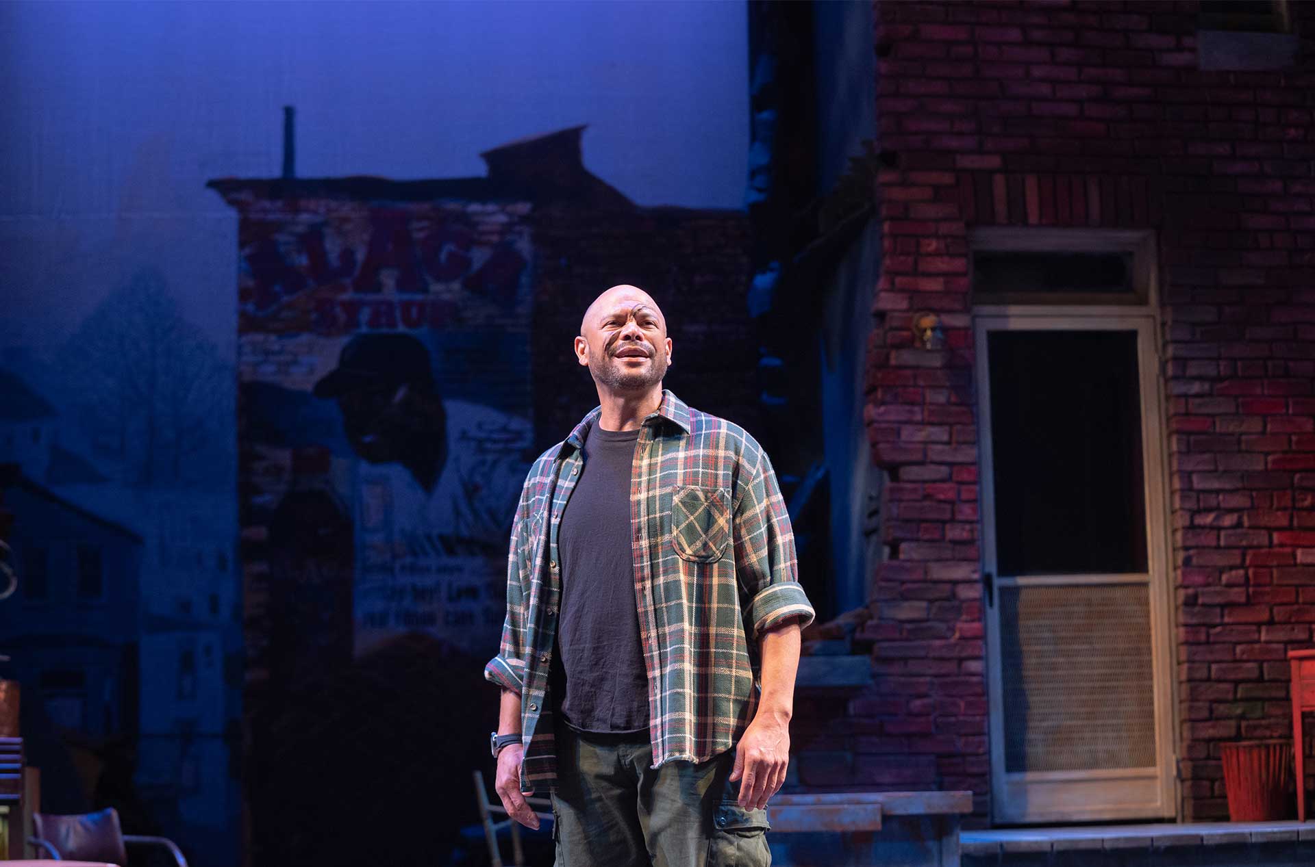 A performer stands alone under cool blue lighting with an anguished expression. A brick house facade and painted wall mural fill the background.