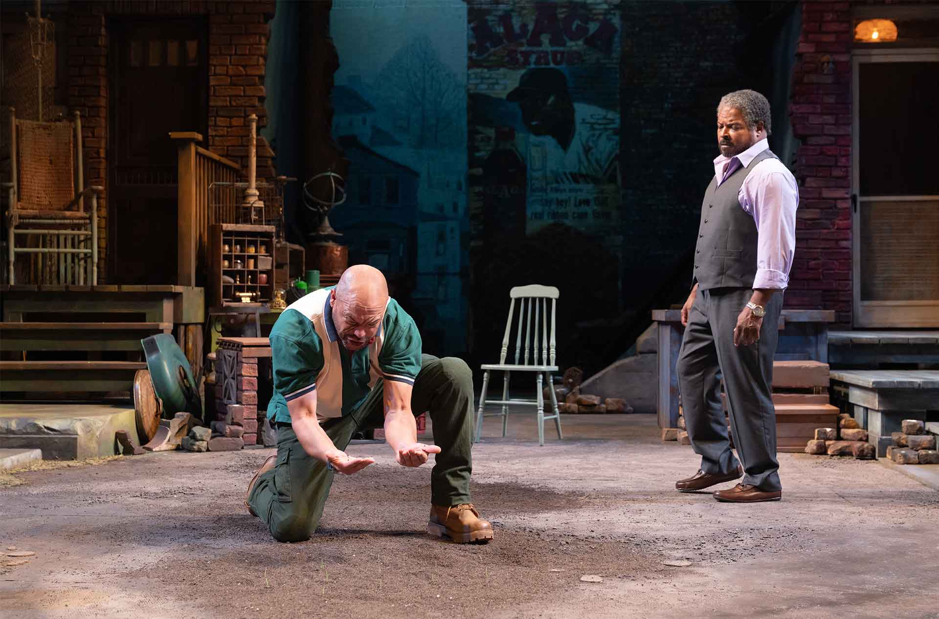 One performer kneels with hands outstretched while another watches from across a worn neighborhood courtyard set. Brick porches and scattered rubble frame the tense scene.