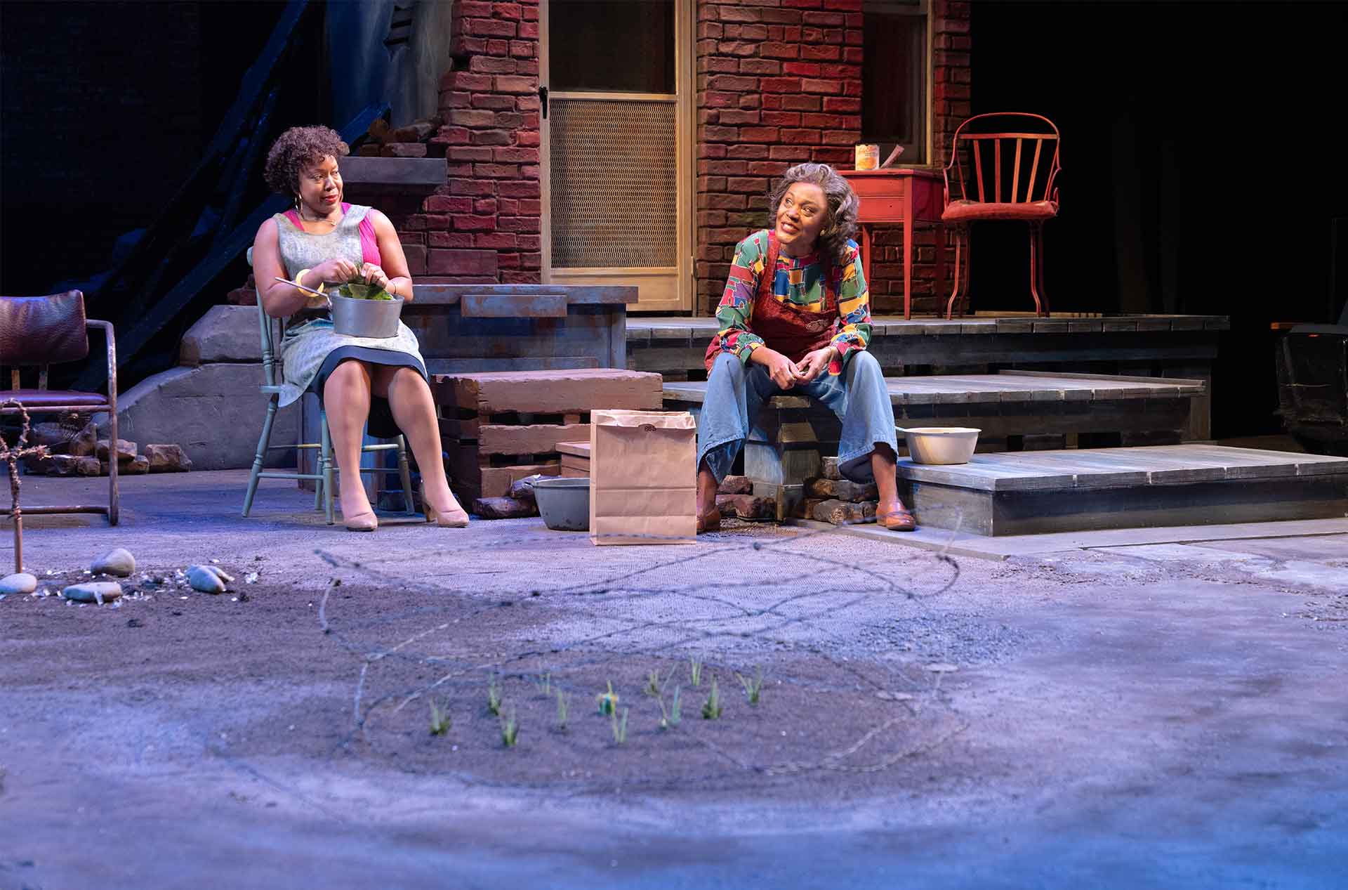Two performers sit outside a brick home while one prepares food in a pot and the other speaks from the steps. Small plants grow from a circle drawn in the dirt before them.