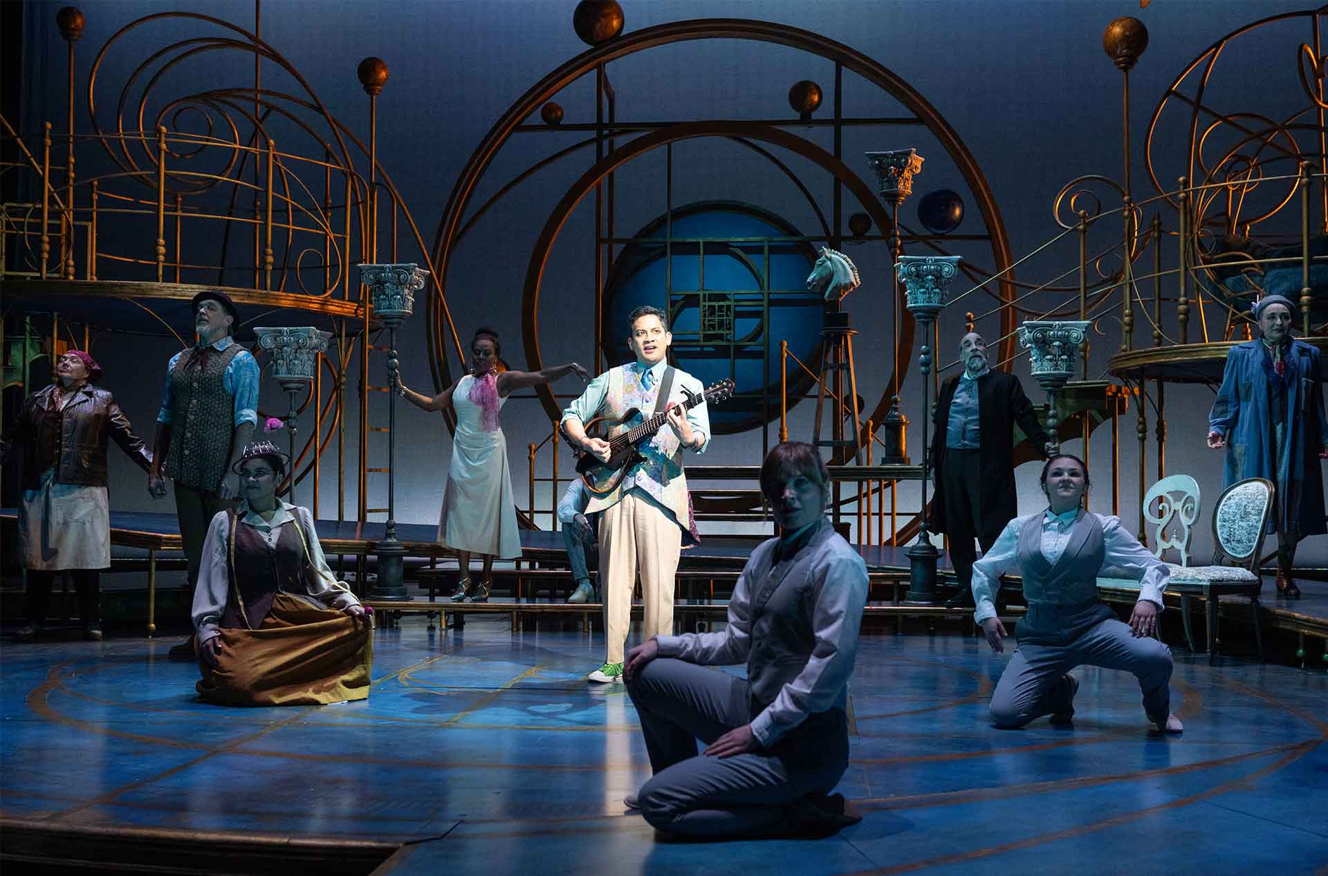 Ensemble cast arranged across a stage with a central performer playing guitar, framed by ornate circular set pieces and soft blue lighting.