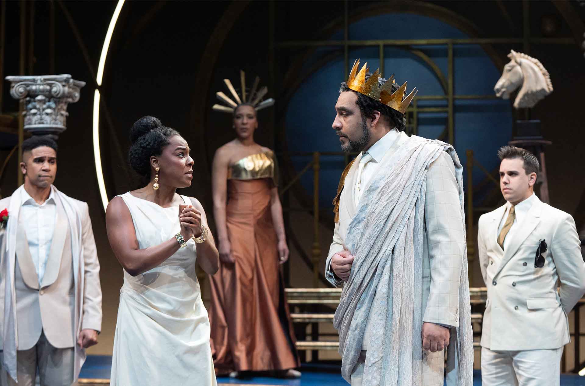 Two performers face each other in an intense moment, one wearing a crown and draped costume, while others stand behind them on a stylized stage.