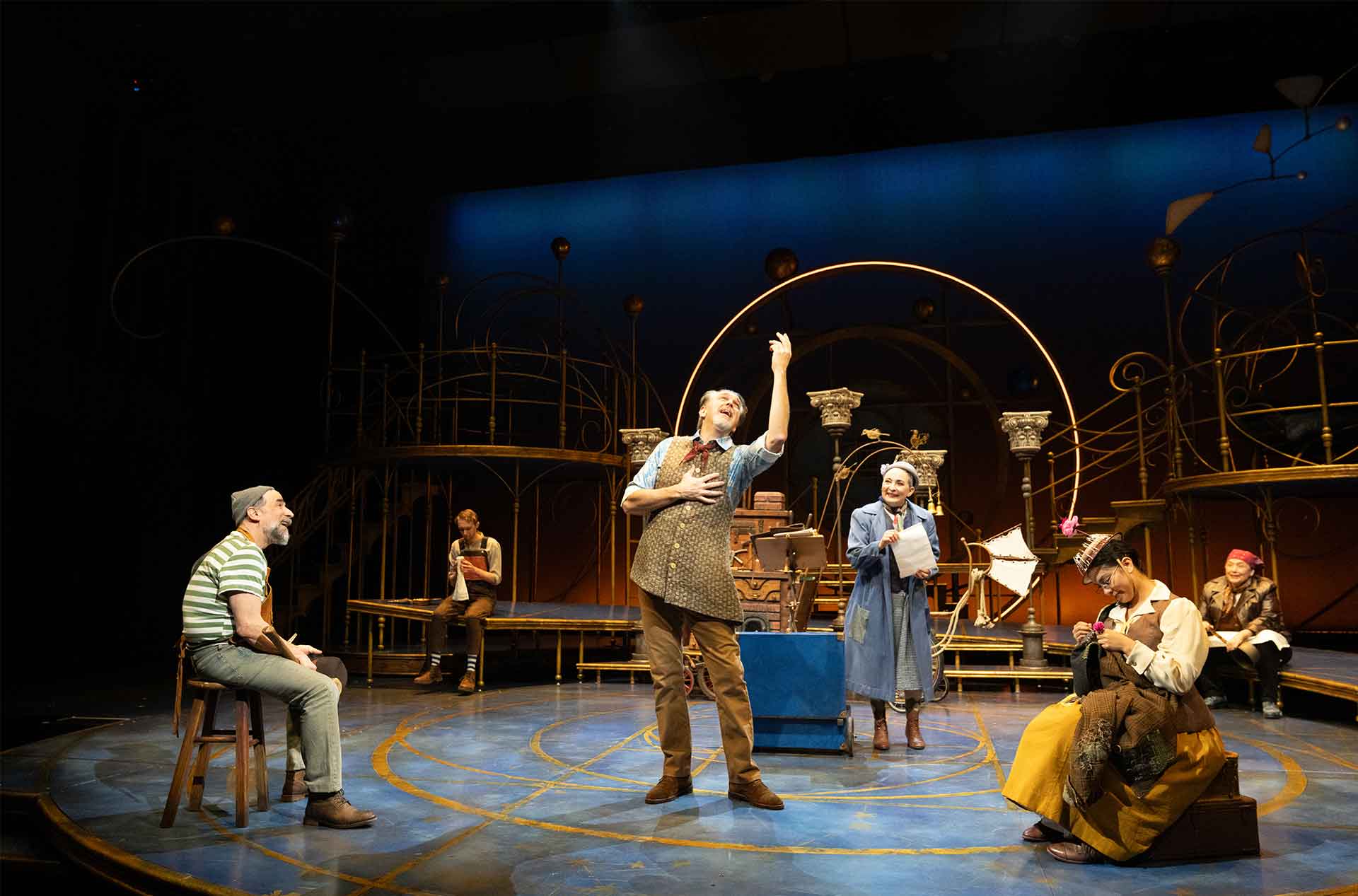 Performer sings expressively at center stage while others sit and stand around in period inspired costumes on a warm lit theatrical set.