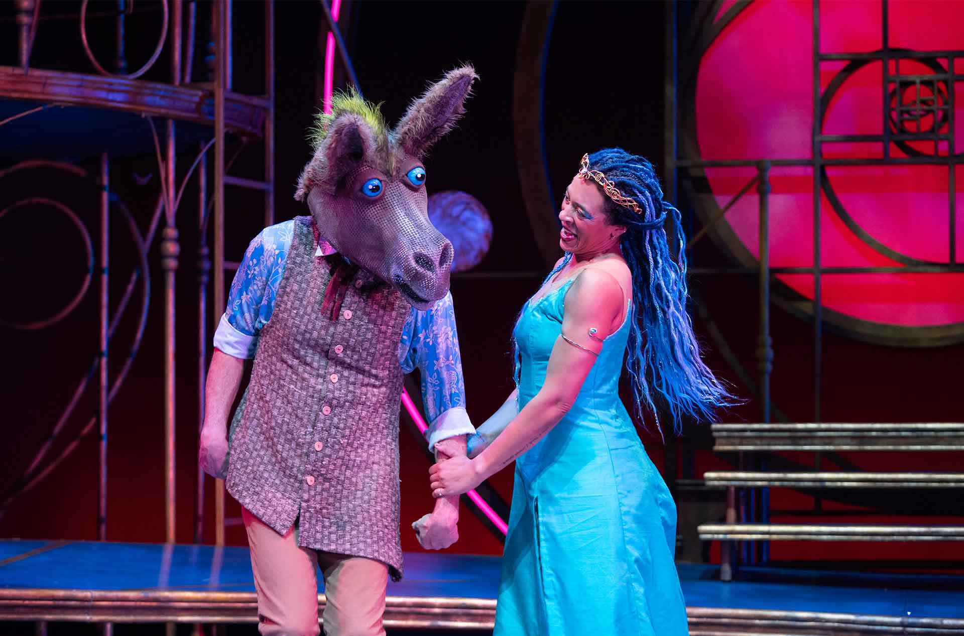 Performer wearing a donkey head mask holds hands with another performer in a blue dress, both smiling under vibrant stage lighting.