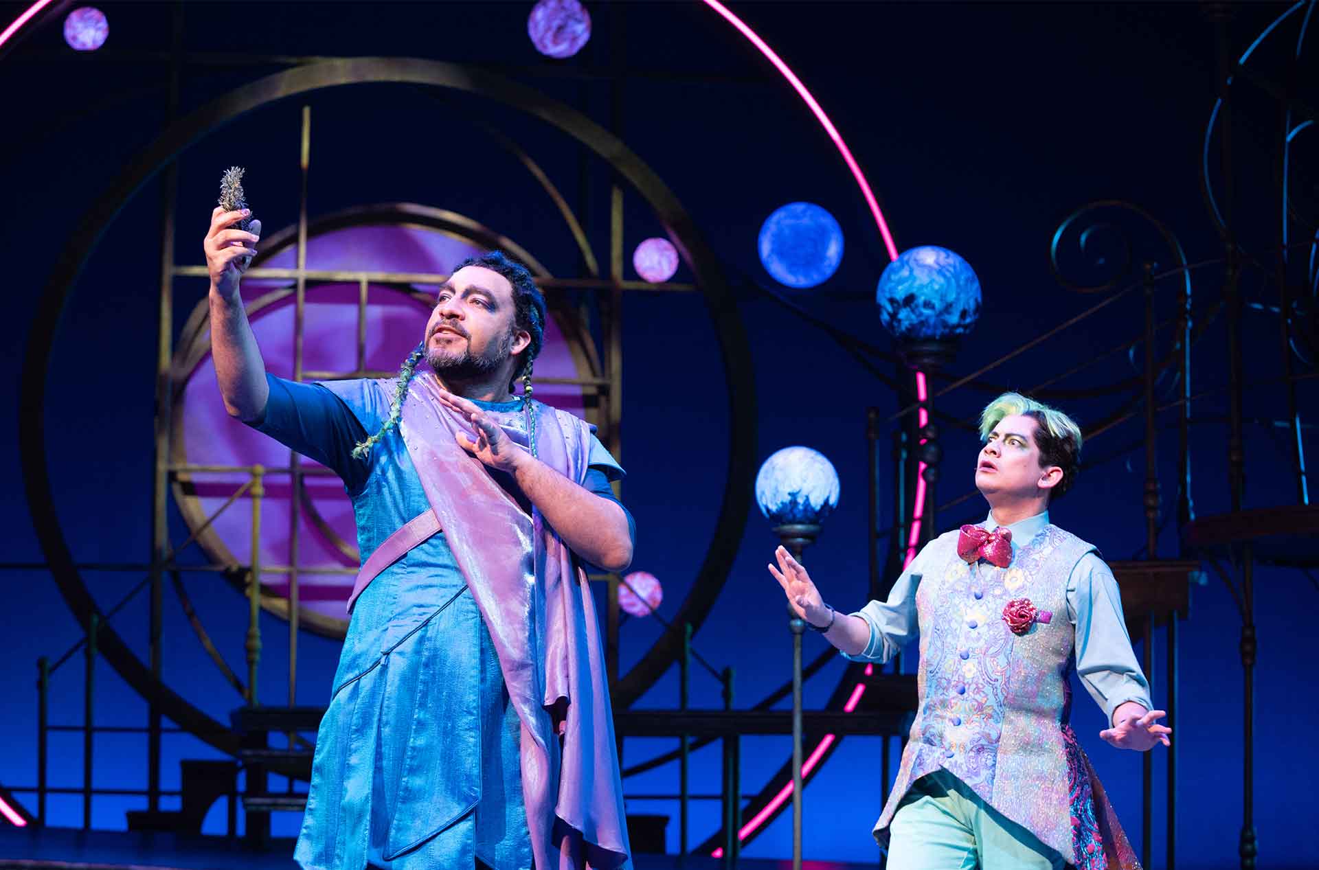 Two performers gesture dramatically, one holding a small object up to the light, against a backdrop of circular set elements and glowing orbs.