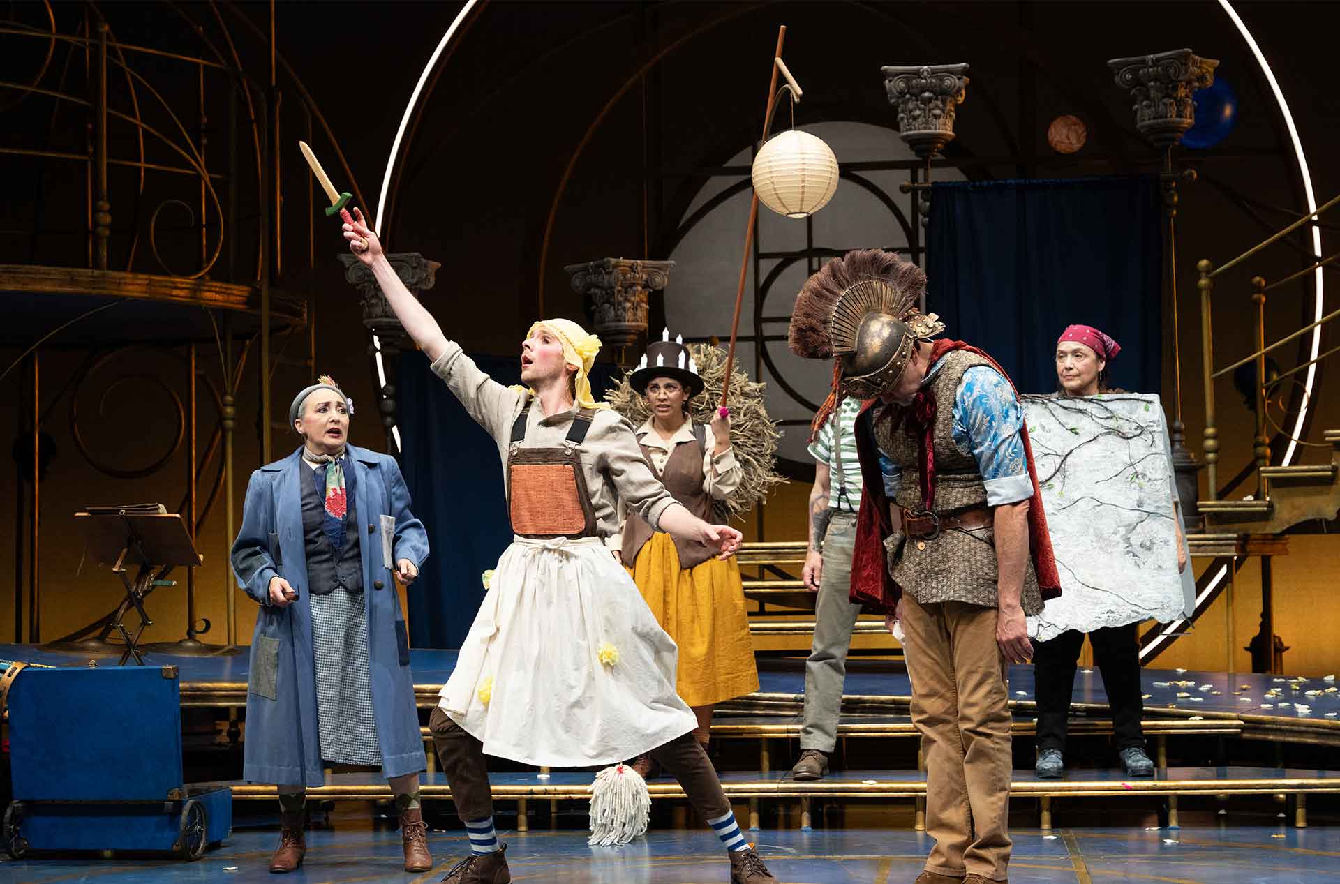 Performer in apron and headscarf raises a prop sword dramatically while others in varied costumes react around them on stage.
