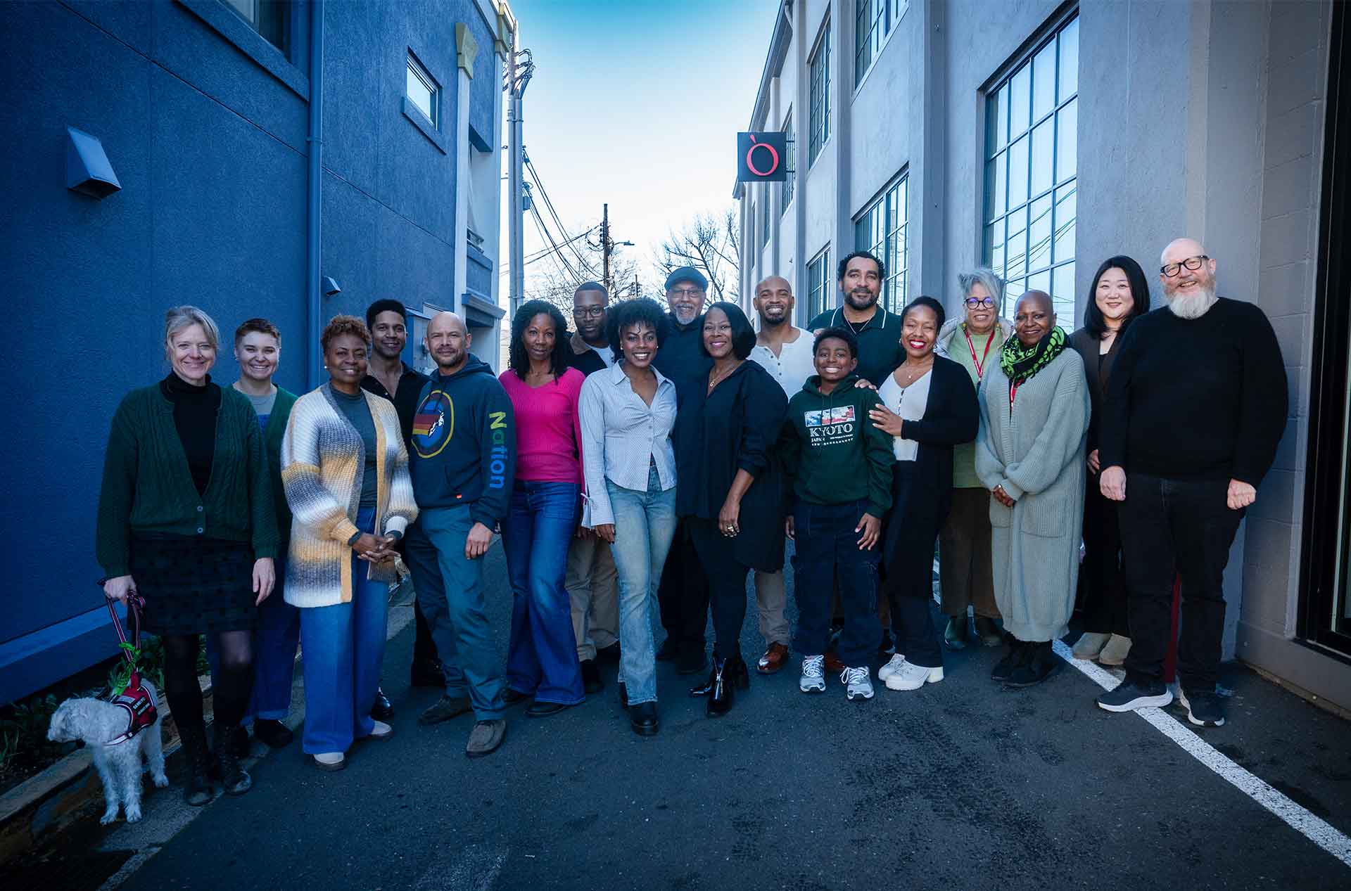 Cast and crew posing together outdoors between two buildings, smiling in front of the theater’s logo.