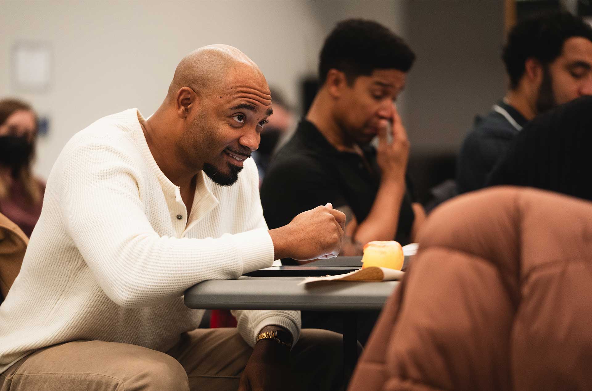 Person smiling and gesturing while eating, with others around the table focused on their scripts.