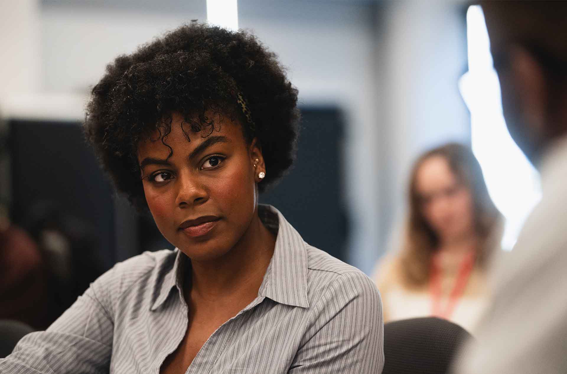 Person with curly hair and striped shirt listening intently during a discussion, with blurred figures in the background.