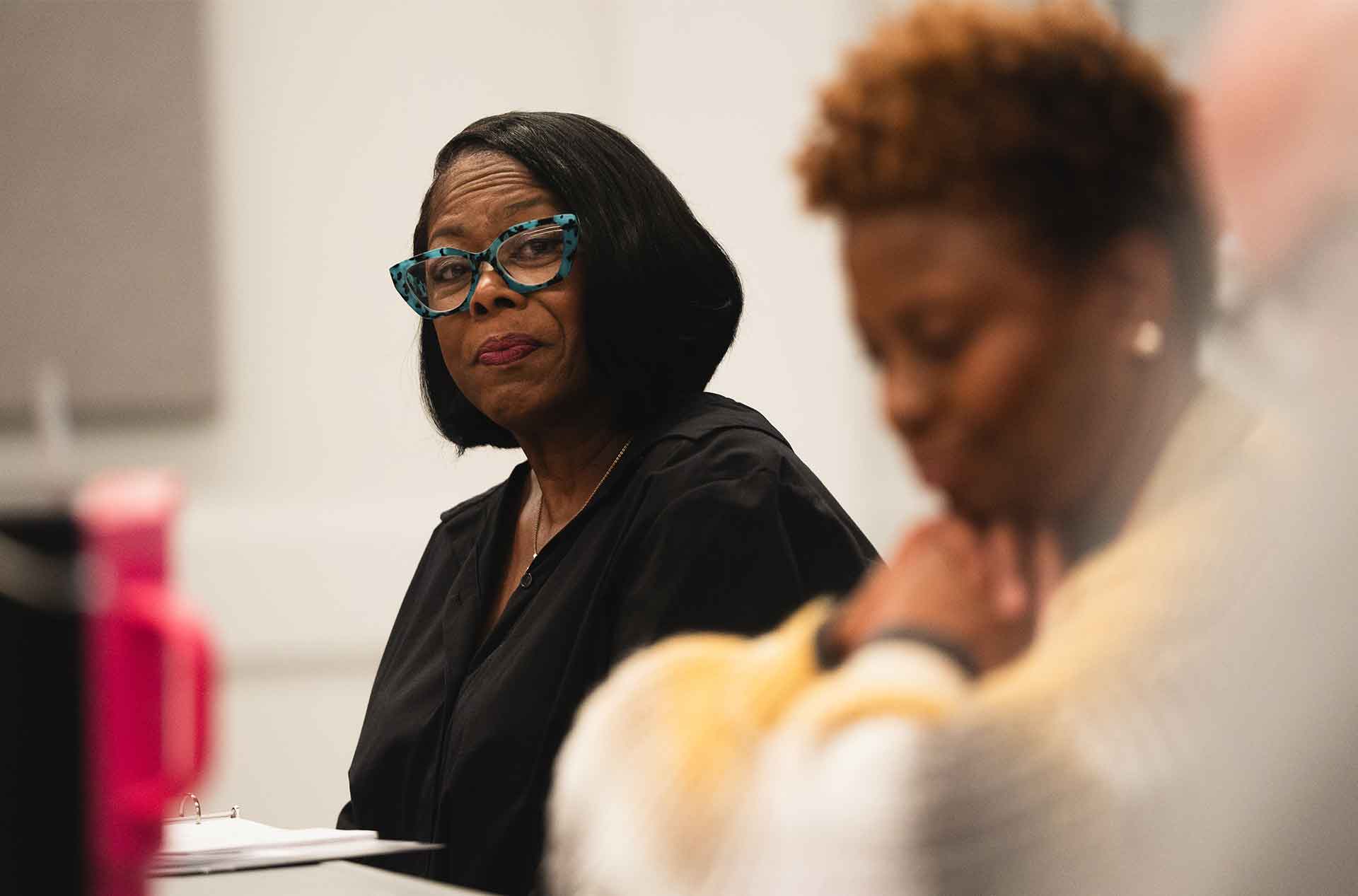 Person with teal glasses listening attentively while another participant is blurred in the foreground.