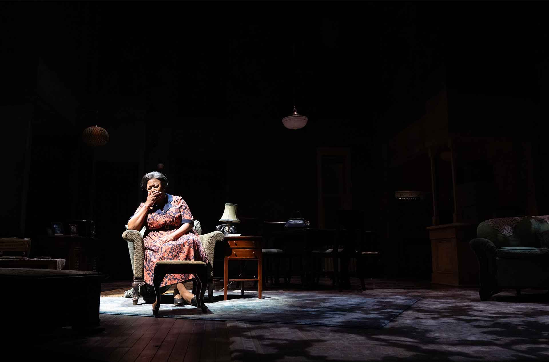 A person sits alone in a dimly lit room with head resting on hand, conveying a quiet or reflective moment.