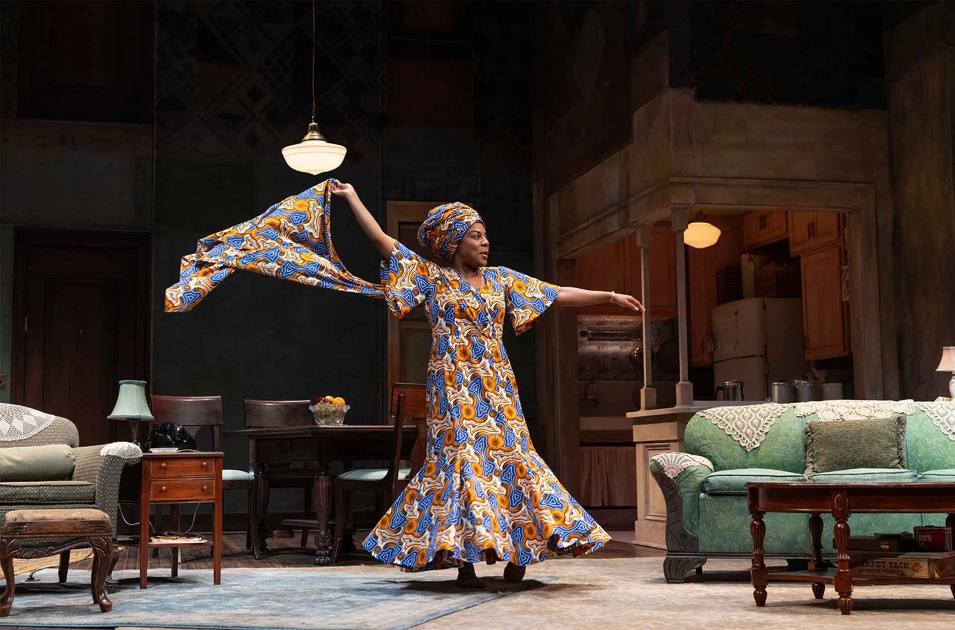 A person in a flowing patterned dress dances across a living room, holding fabric outstretched mid motion.