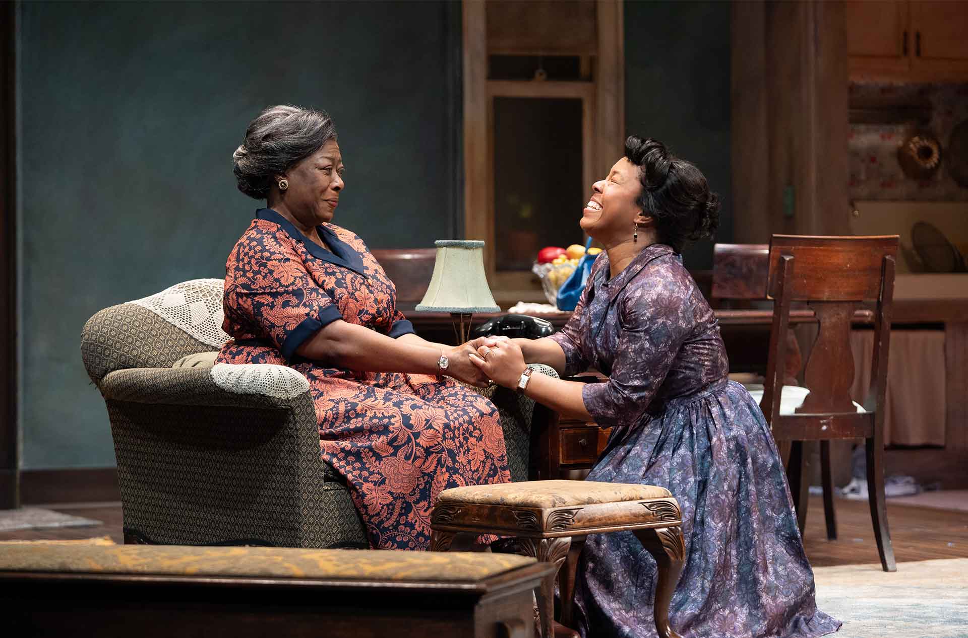 Two people hold hands in a living room while one sits and the other kneels, expressing an emotional moment.