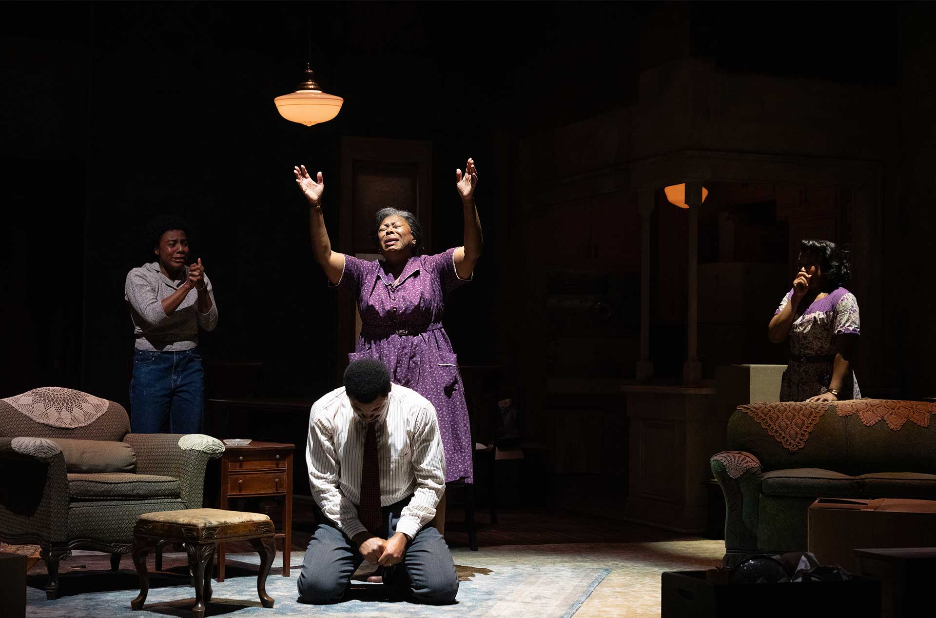 One person kneels with head bowed while another stands with arms raised under a single overhead light, others nearby reacting.