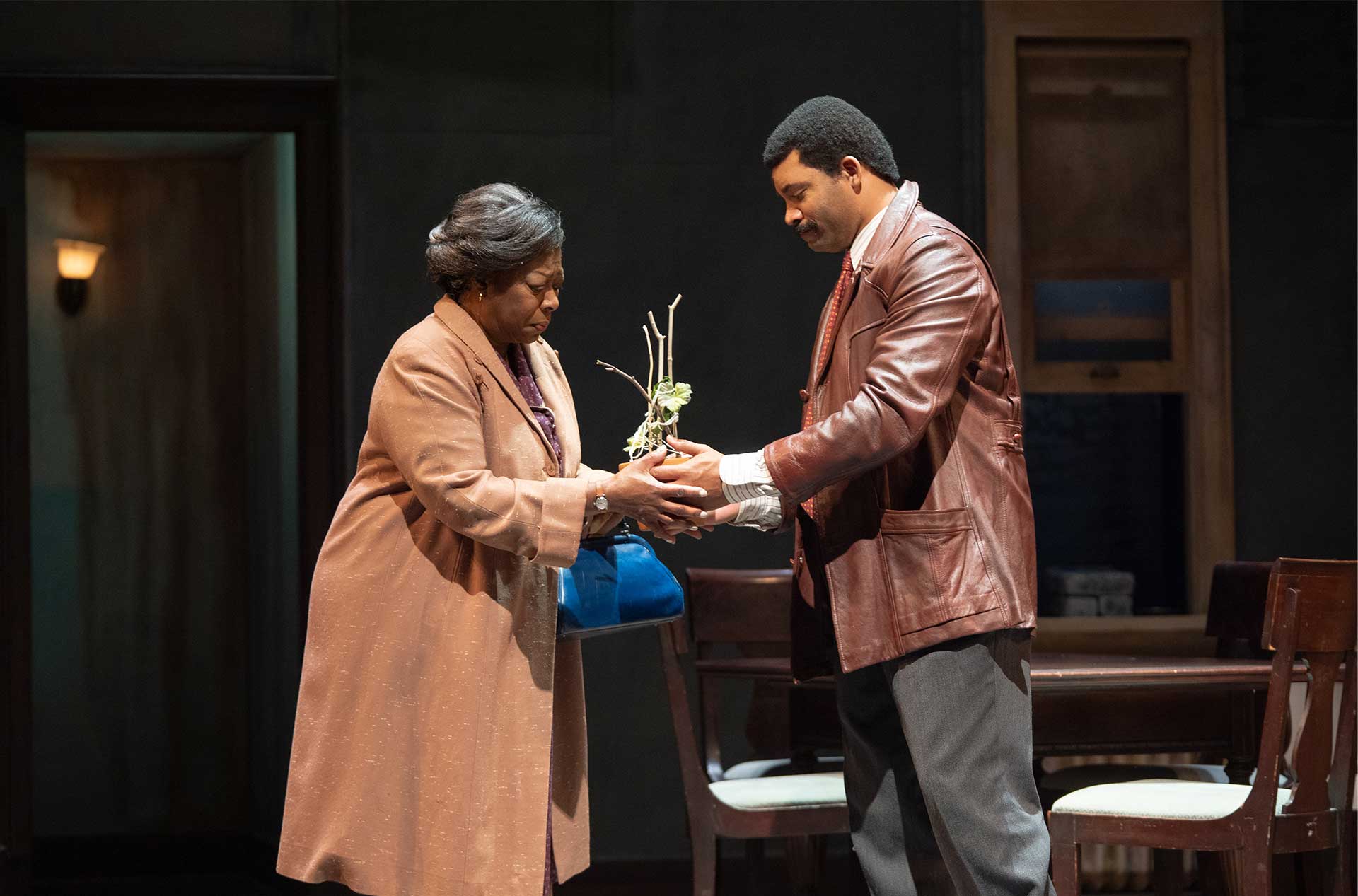 One person hands a small plant to another in a living room, suggesting a meaningful exchange.