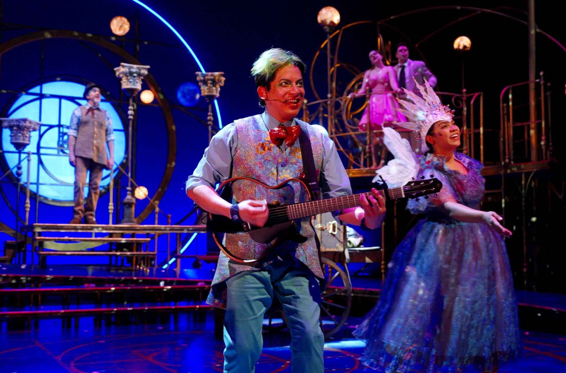 Performer with short dyed hair plays a small guitar and smiles at the audience on a brightly lit stage with colorful costumes and whimsical set pieces while other cast members sing and dance in the background.