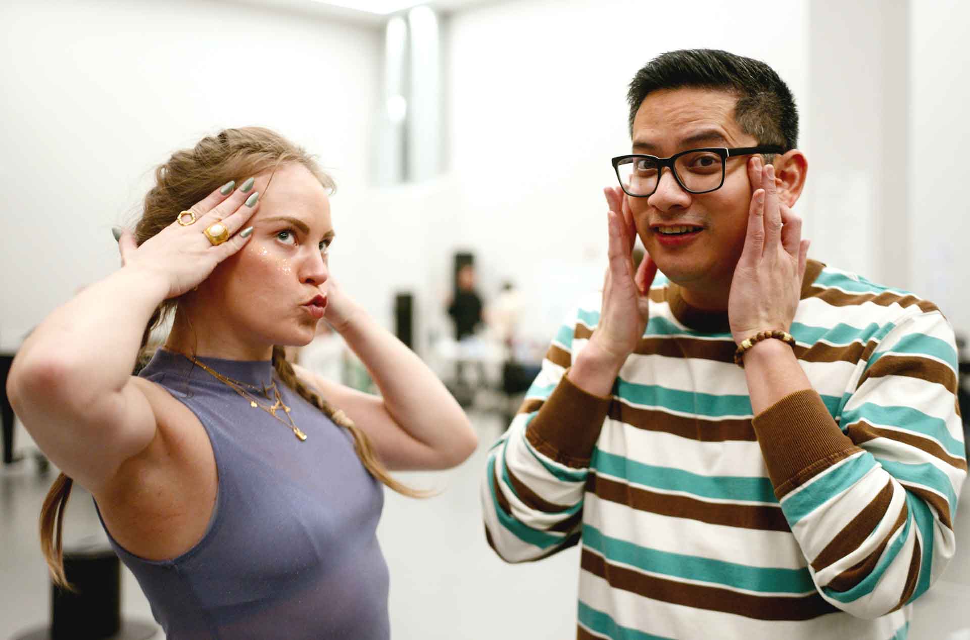 Two people stand indoors touching their cheeks with both hands as if reacting or practicing facial expressions. One wears glasses and a striped sweater while the other wears a sleeveless top and jewelry in a bright studio setting.
