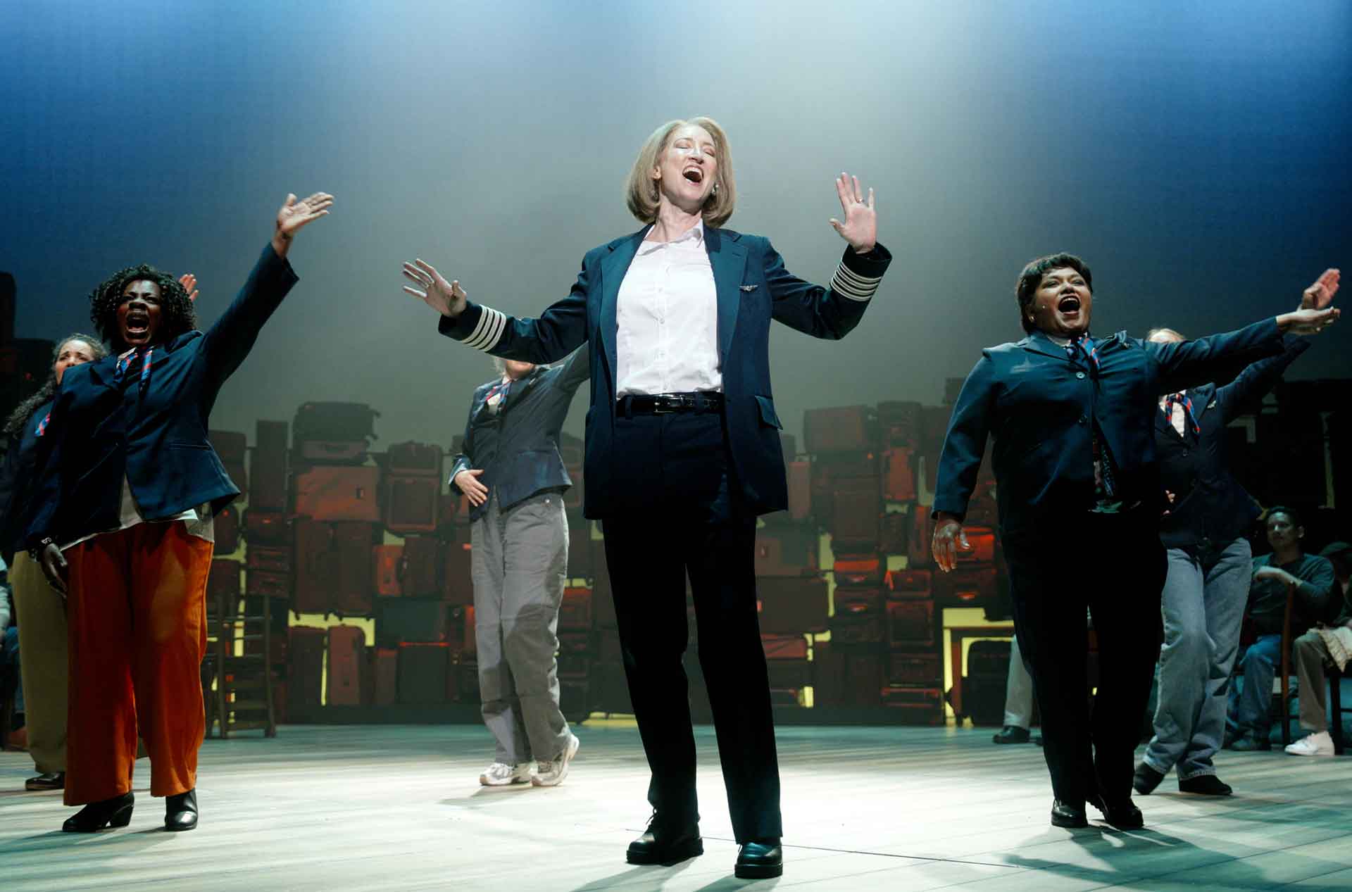 Group of performers on a stage mid-song with arms raised and expressive faces under soft theatrical lighting.
