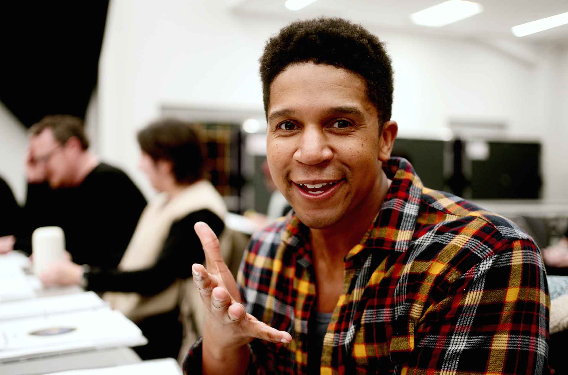 Person in a red and yellow plaid shirt smiling and gesturing with one hand while seated at a table in a classroom setting, with others blurred in the background.