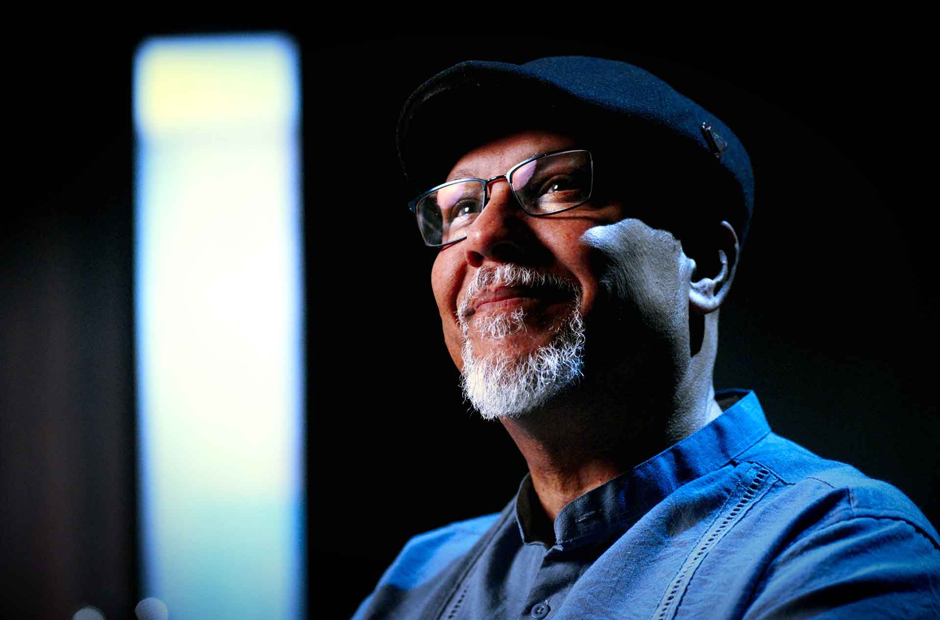 Portrait of a person with a white beard wearing glasses and a dark flat cap. The person looks slightly upward with a soft smile against a dark background with a vertical strip of light beside them.