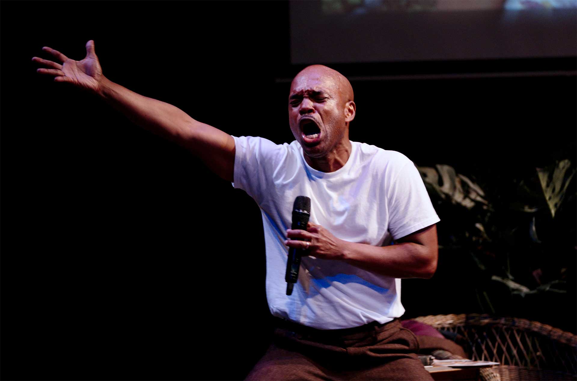 Person on stage holding a microphone and extending one arm during a dramatic performance.