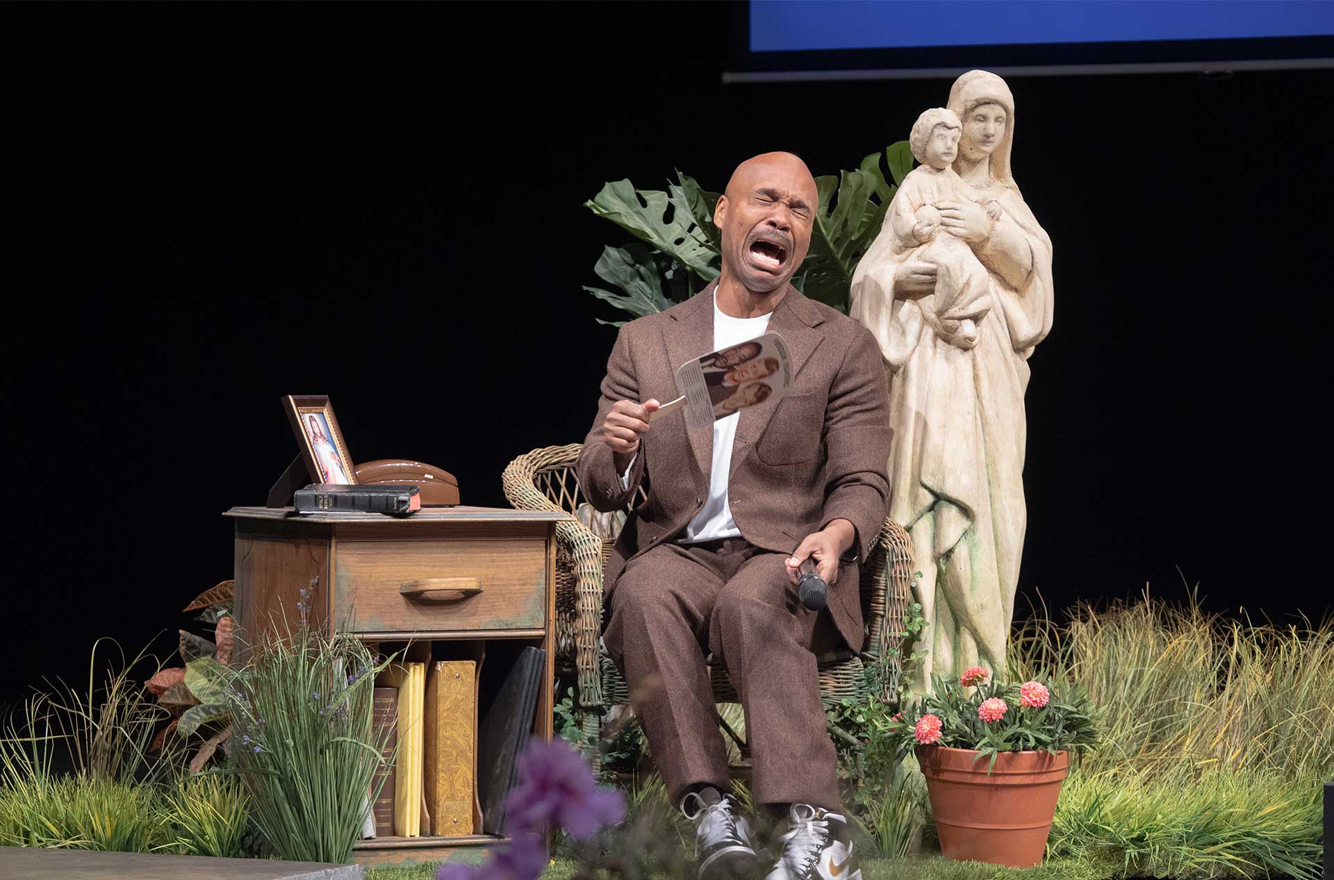 Performer sits beside a religious statue and plants while holding a microphone and reacting intensely.