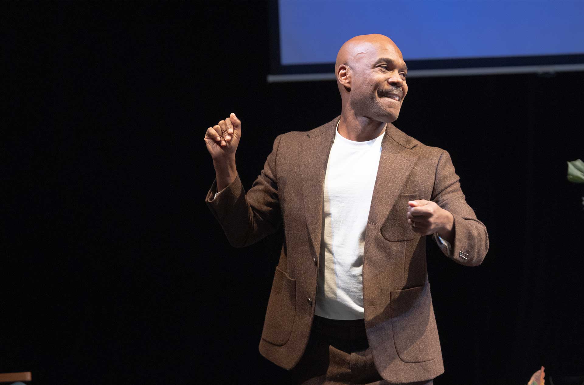 Performer smiles and gestures onstage in a brown suit jacket against a dark backdrop.