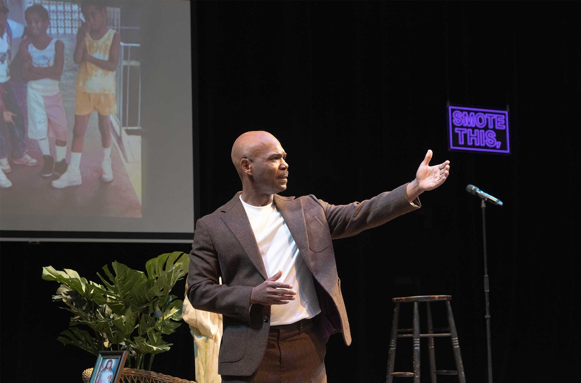 Performer gestures outward beside a microphone and stool while a projected childhood photo appears behind.