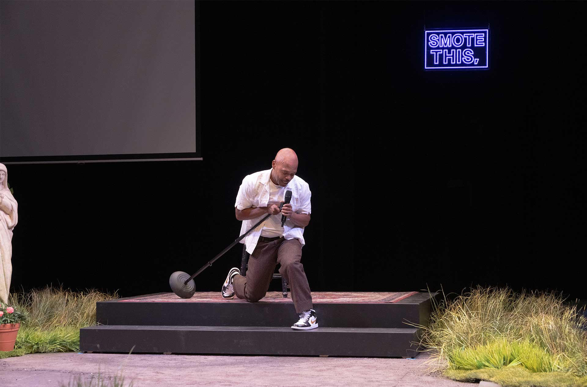 Performer kneels onstage holding a microphone beneath a glowing sign that reads "SMOTE THIS."