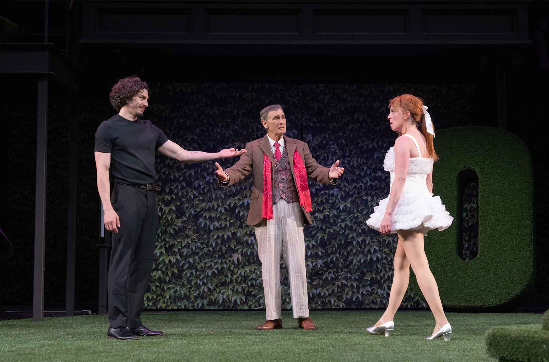 Three performers face each other on a turf covered stage. One wears a white ruffled dress while the others gesture toward them in conversation.