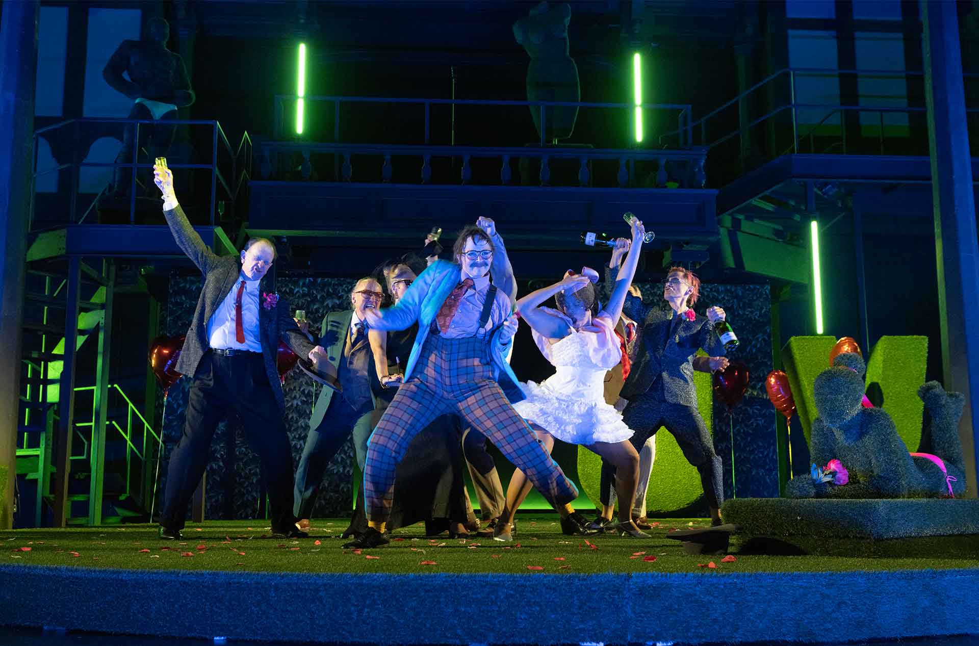A large ensemble celebrates on a green stage under bright neon lighting. Performers raise glasses and bottles while flower petals scatter across the floor.