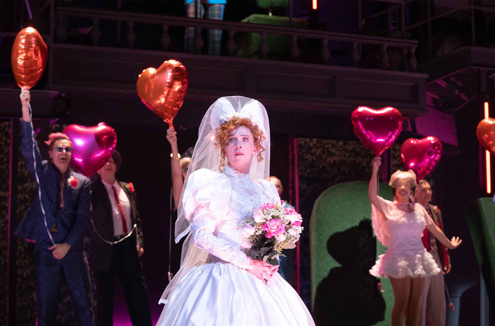 A performer in a white wedding dress and veil holds a bouquet at center stage. Other performers behind them hold red and pink heart shaped balloons.