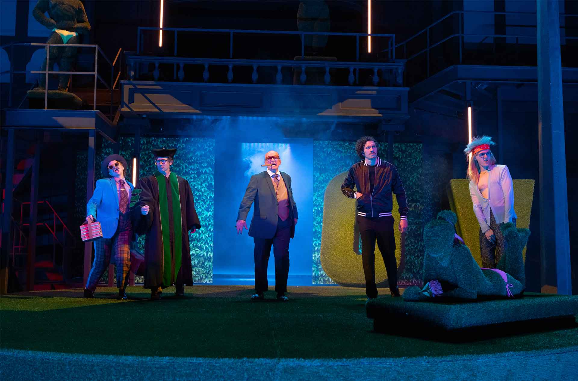 Performers stand across a blue lit stage with smoke in the center doorway. The set includes tall hedges. balcony railings. neon light bars. and a topiary statue.