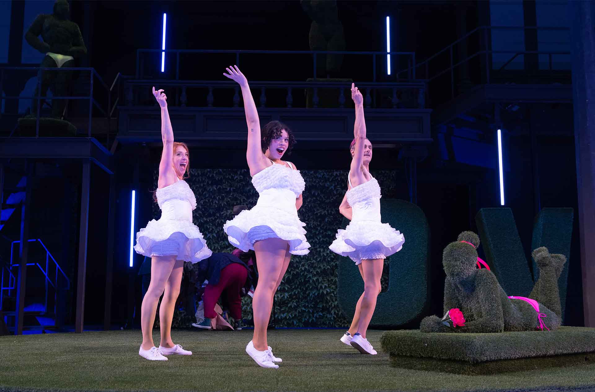 Three performers in white ruffled dresses dance with raised arms on a neon lit stage. A topiary statue sits beside them on a raised turf platform.