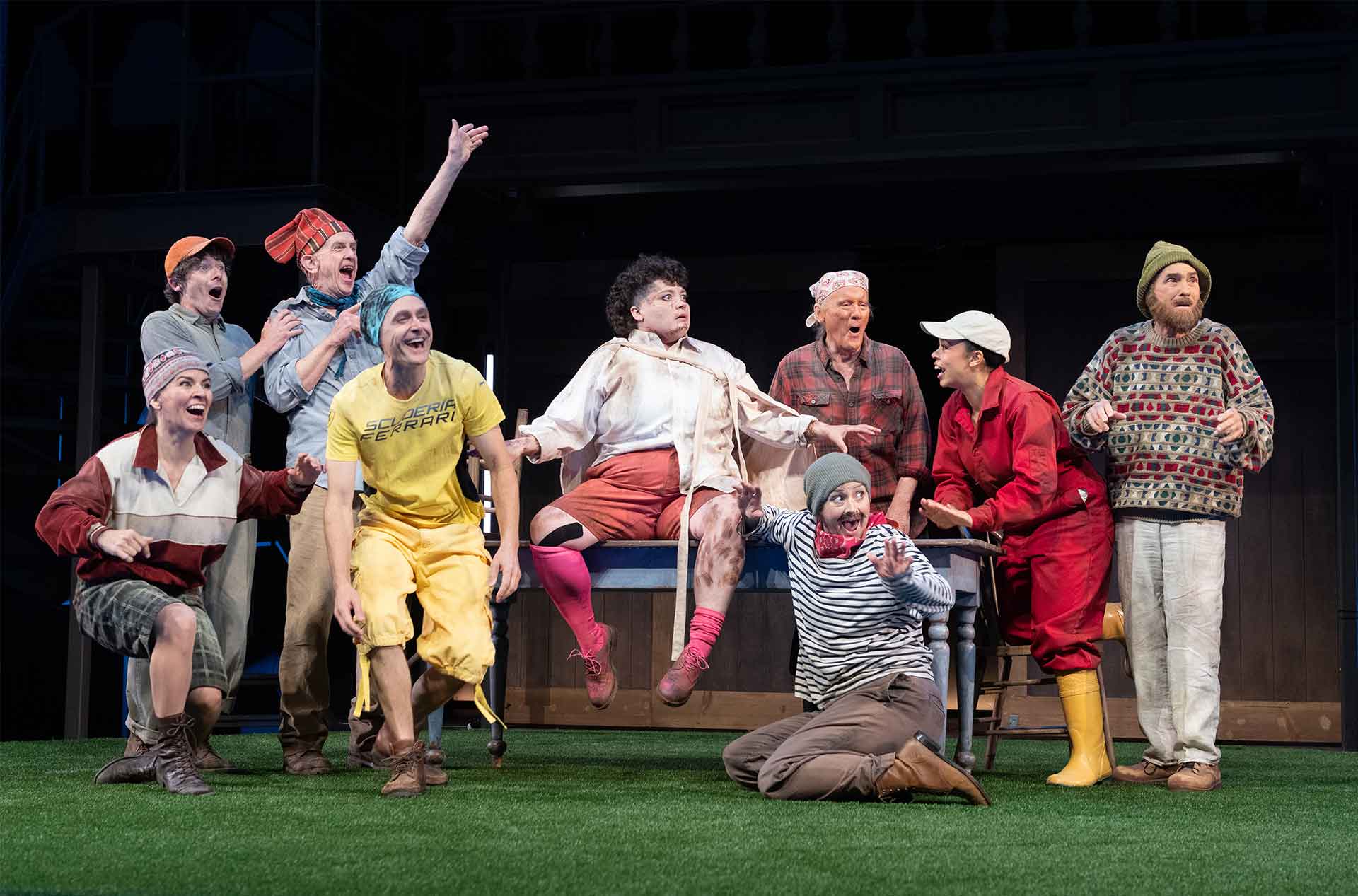 A large ensemble in colorful casual costumes gathers around a table on a turf covered stage.