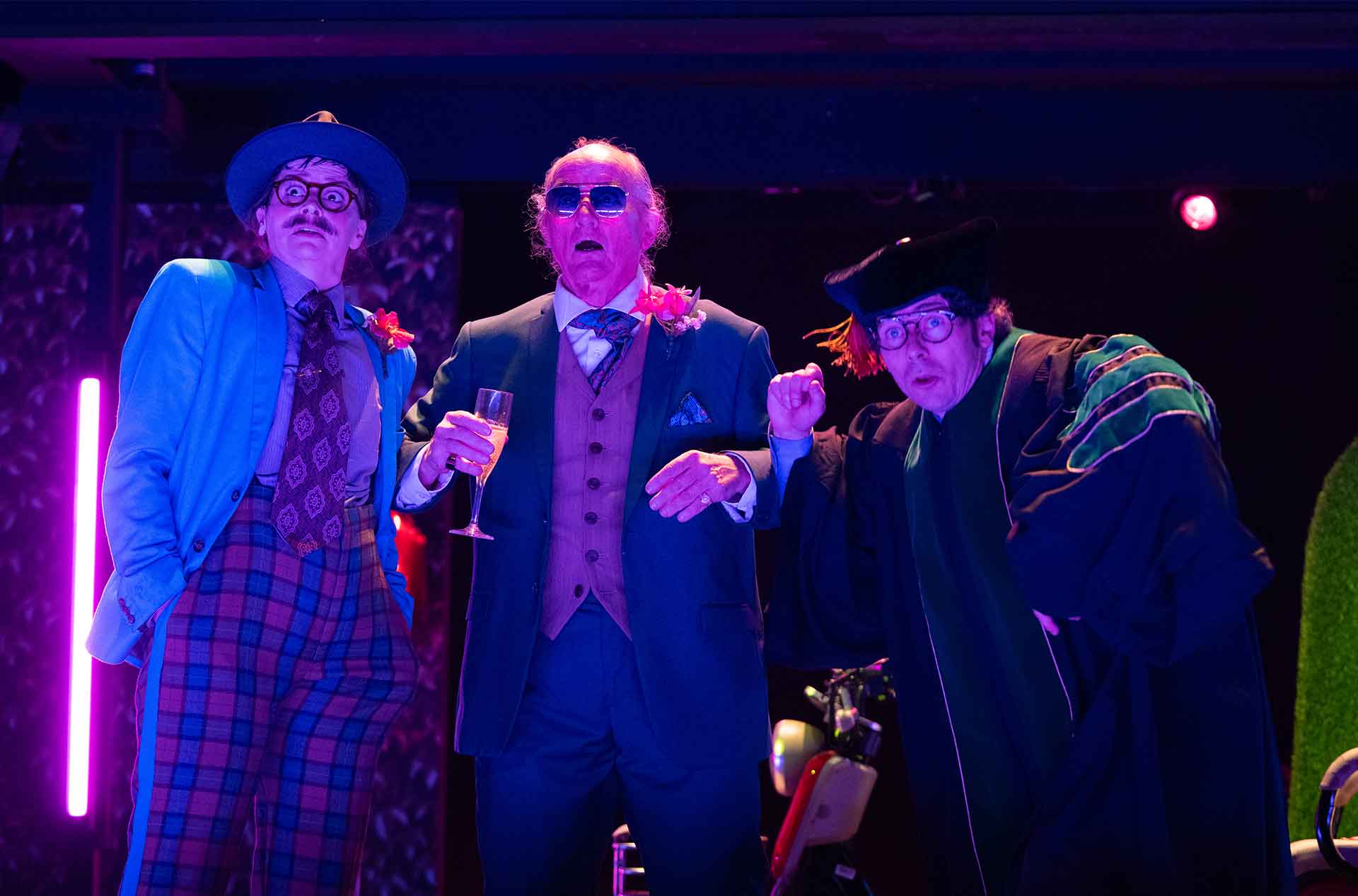 Three performers in formal and academic costumes lean toward the audience with surprised expressions. Purple and blue stage lighting frames them from behind.