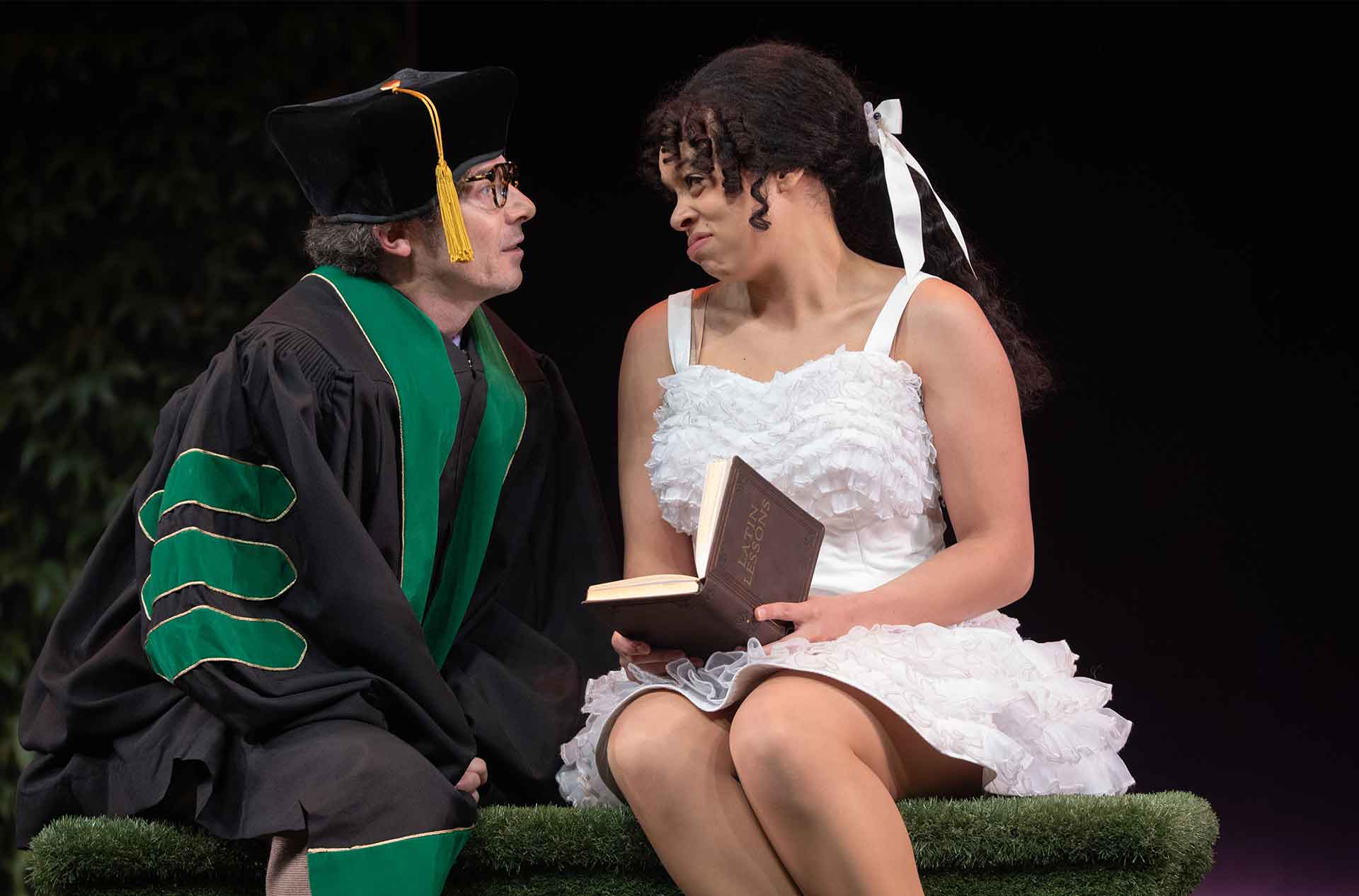 A performer in academic robes leans toward a seated performer in a white ruffled dress holding a book labeled "Life Lessons". They exchange an intense look on a dark stage.