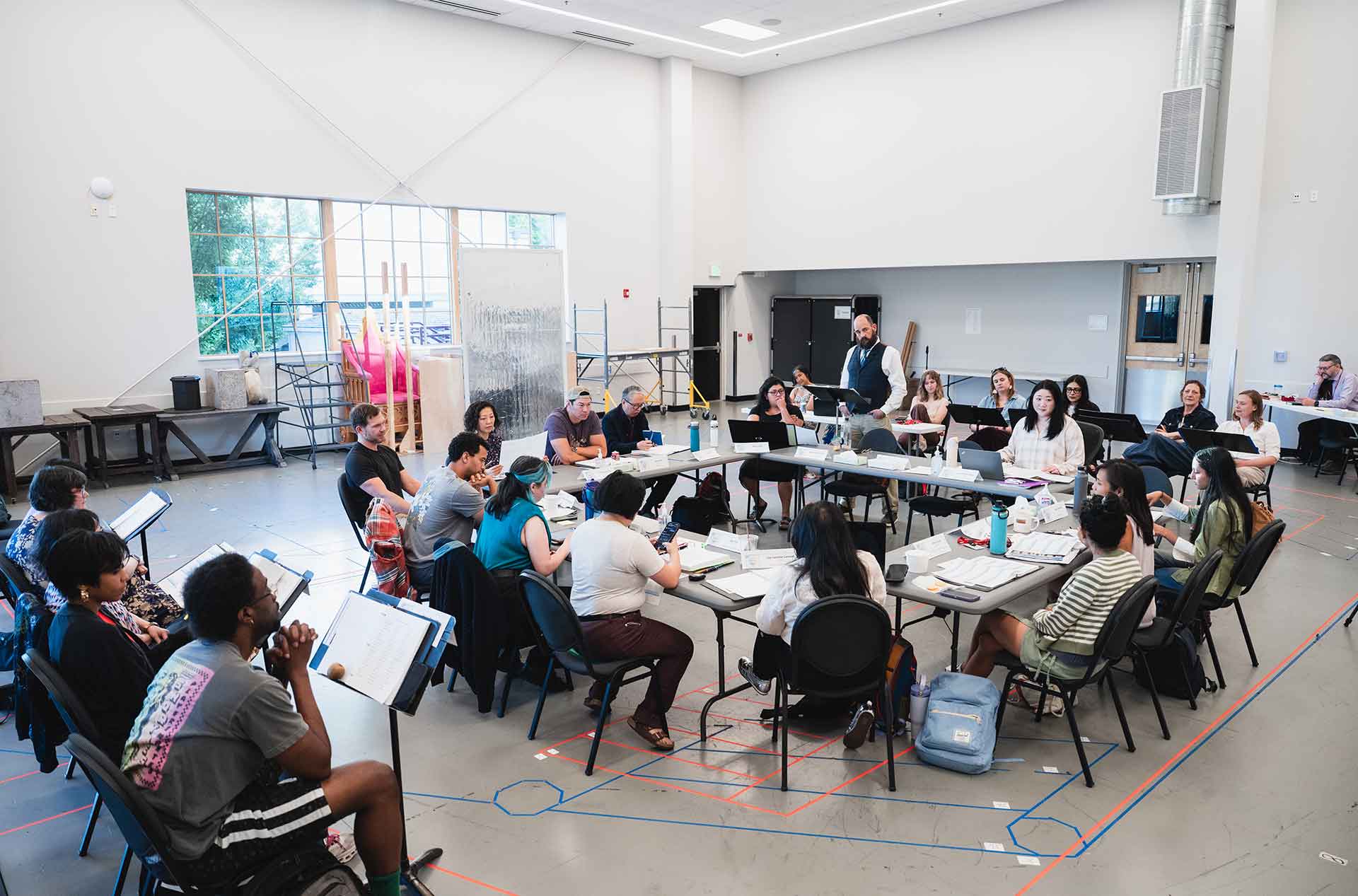 Wide view of a play rehearsal with cast members seated around tables in a rehearsal room.