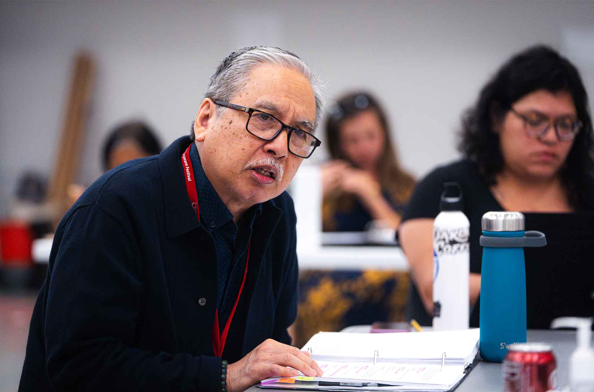 Person with glasses reviews a rehearsal binder while seated at a table with others in the background.