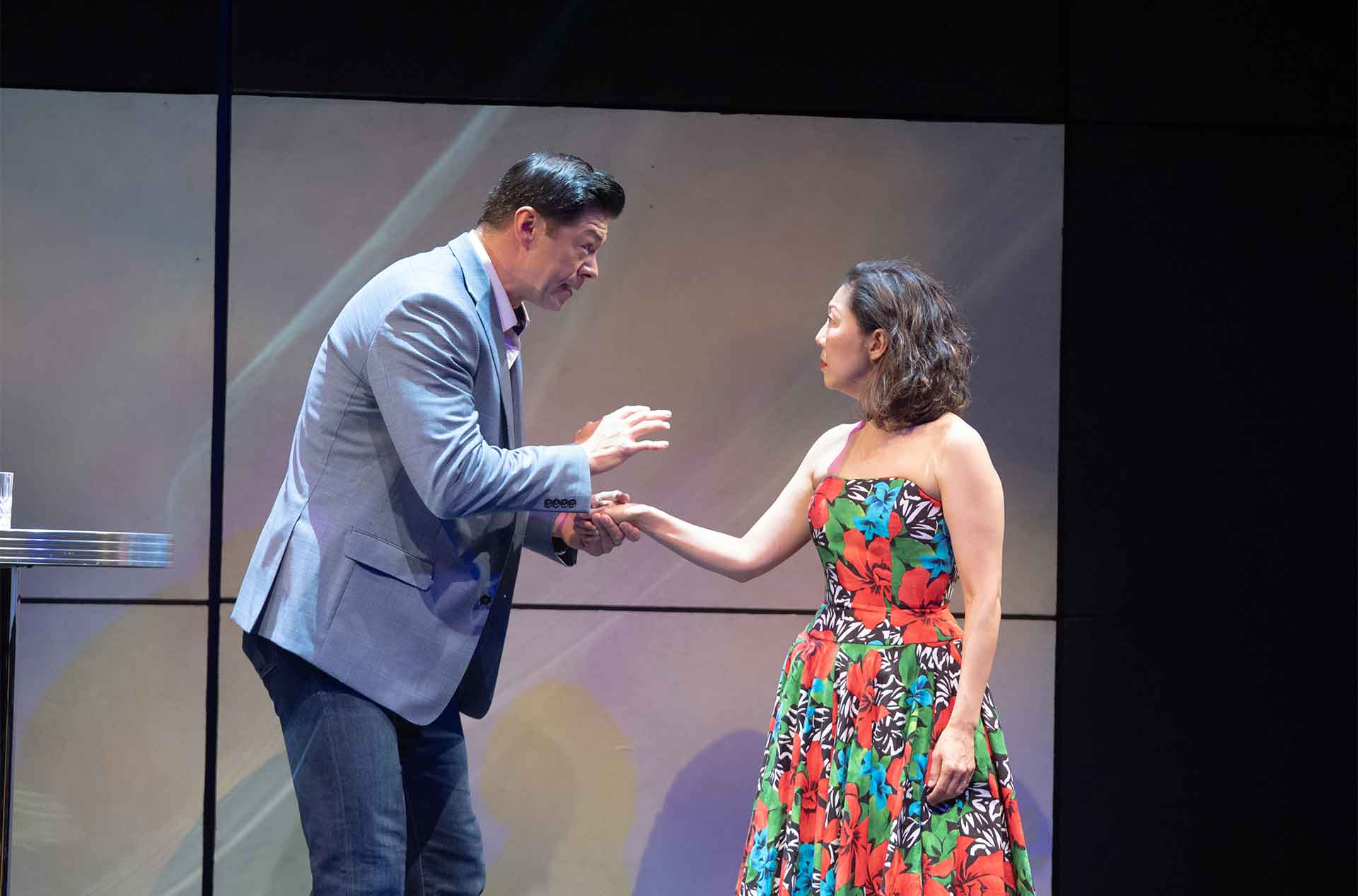 A man in a blue blazer gently holds the hand of a woman wearing a colorful floral dress as they face each other on stage. Their close posture and serious expressions suggest an intimate or emotional conversation.