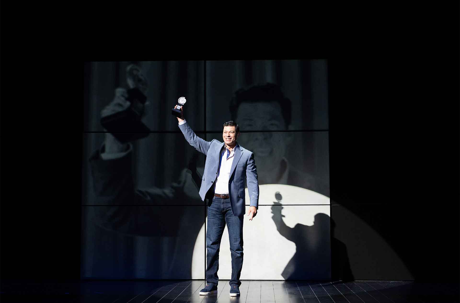 A smiling man in a blue blazer stands in a spotlight and raises an award above his head. A large projection of his face and the raised award fills the stage backdrop behind him.