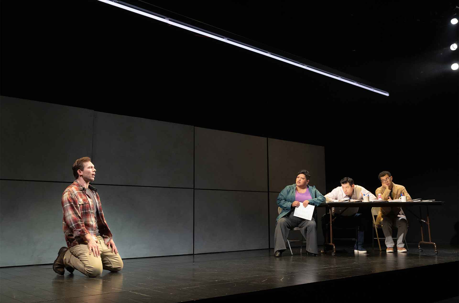 Alt text: A man kneels alone on a brightly lit stage while three people seated at a long table watch him closely. The sparse set and their focused expressions give the scene the feeling of an audition or questioning.