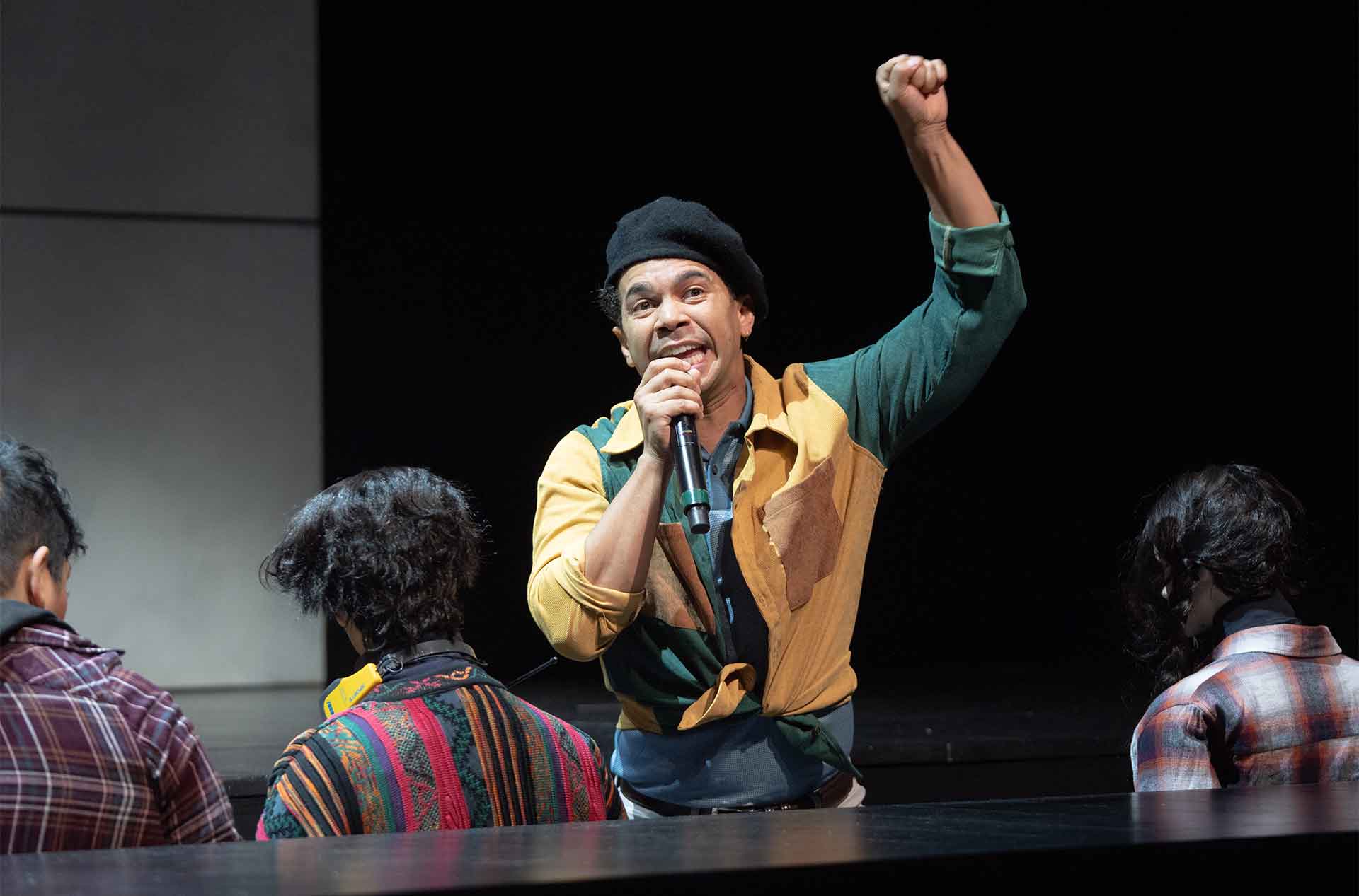 A man wearing a black beret and a multicolored shirt speaks intensely into a microphone while raising one fist above his head. Three seated people are seen from behind in the foreground as he addresses them.