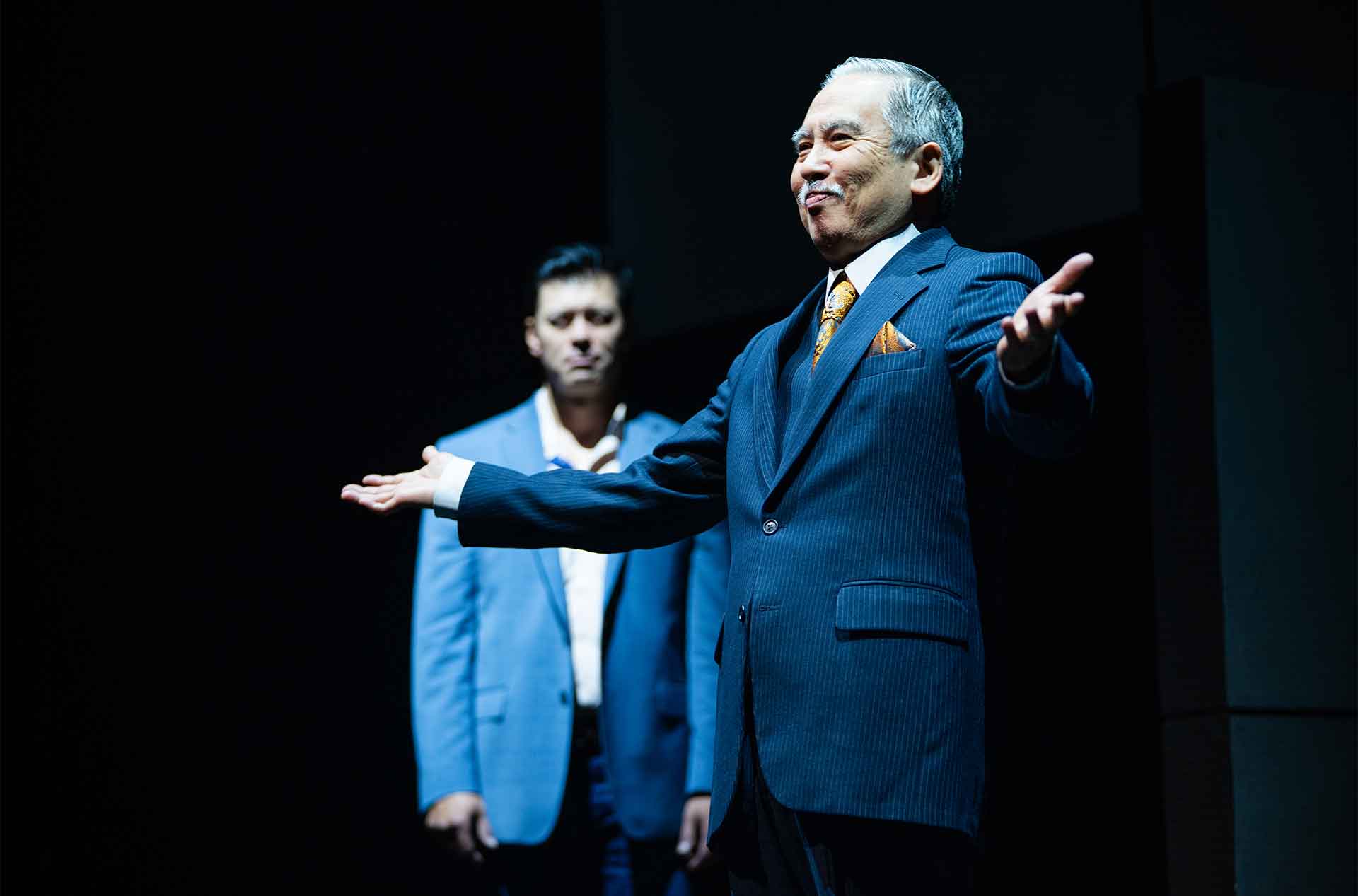 An older man in a dark pinstripe suit smiles and stretches both arms outward under a spotlight. A younger man in a blue blazer stands behind him in shadow watching.