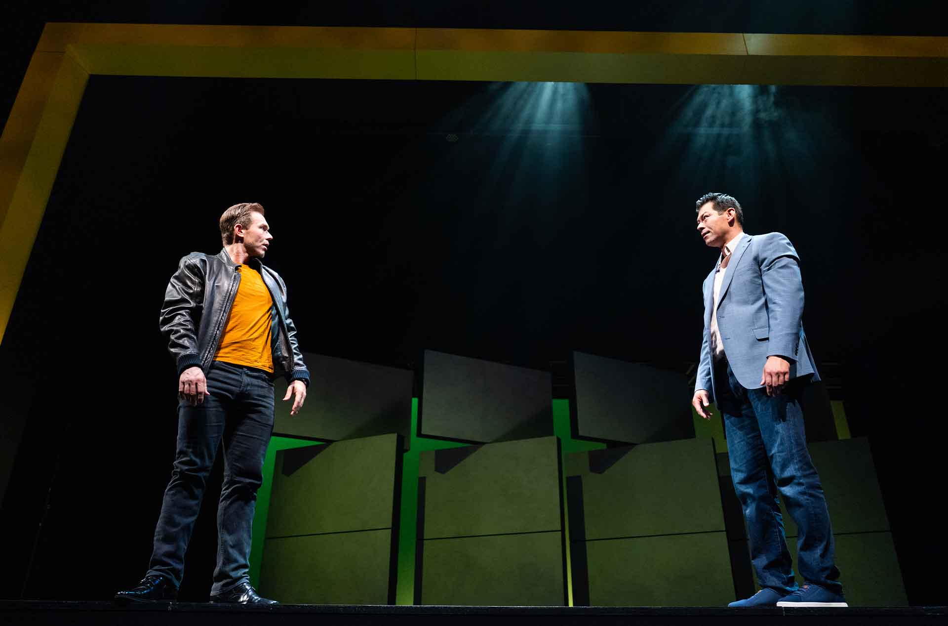 Two men stand far apart facing each other under dramatic overhead stage lighting. One wears a black leather jacket over an orange shirt while the other wears a blue blazer and both appear tense and confrontational.