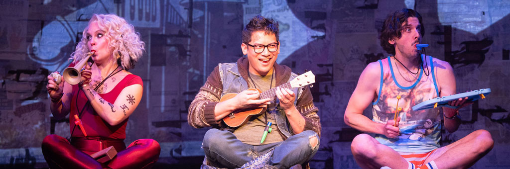 Three performers sit cross legged on a stage playing small toy instruments including a trumpet a ukulele and a xylophone.