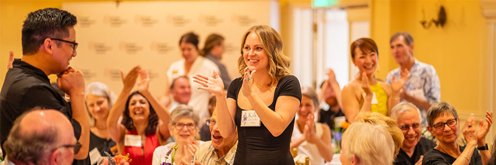 A person standing and smiling is applauded by a room full of seated attendees at a brightly lit celebration or event.