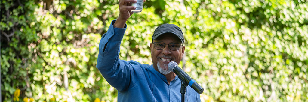 A person wearing glasses and a cap smiles and raises a drink while speaking into a microphone outdoors in front of green foliage.