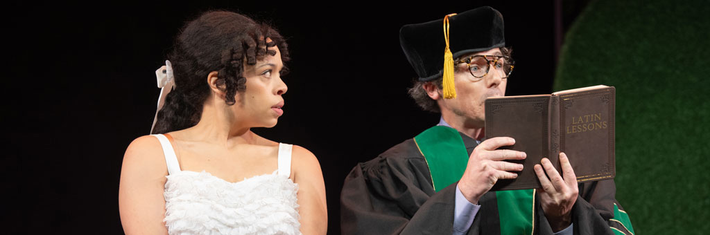 Person in white costume looks to the side while another person in academic robes and cap reads from a book labeled “Latin Lessons” on a dark stage.